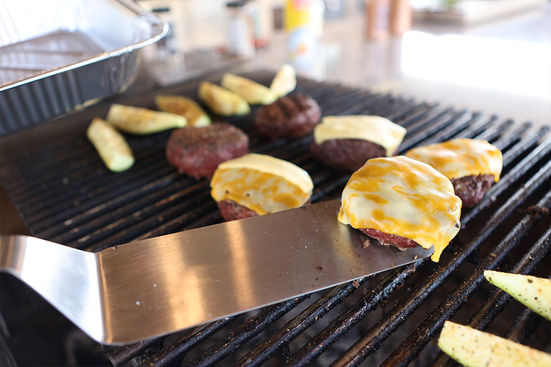 Grilled cheeseburgers and seasoned vegetables cooking on a barbecue, with a spatula lifting one patty.