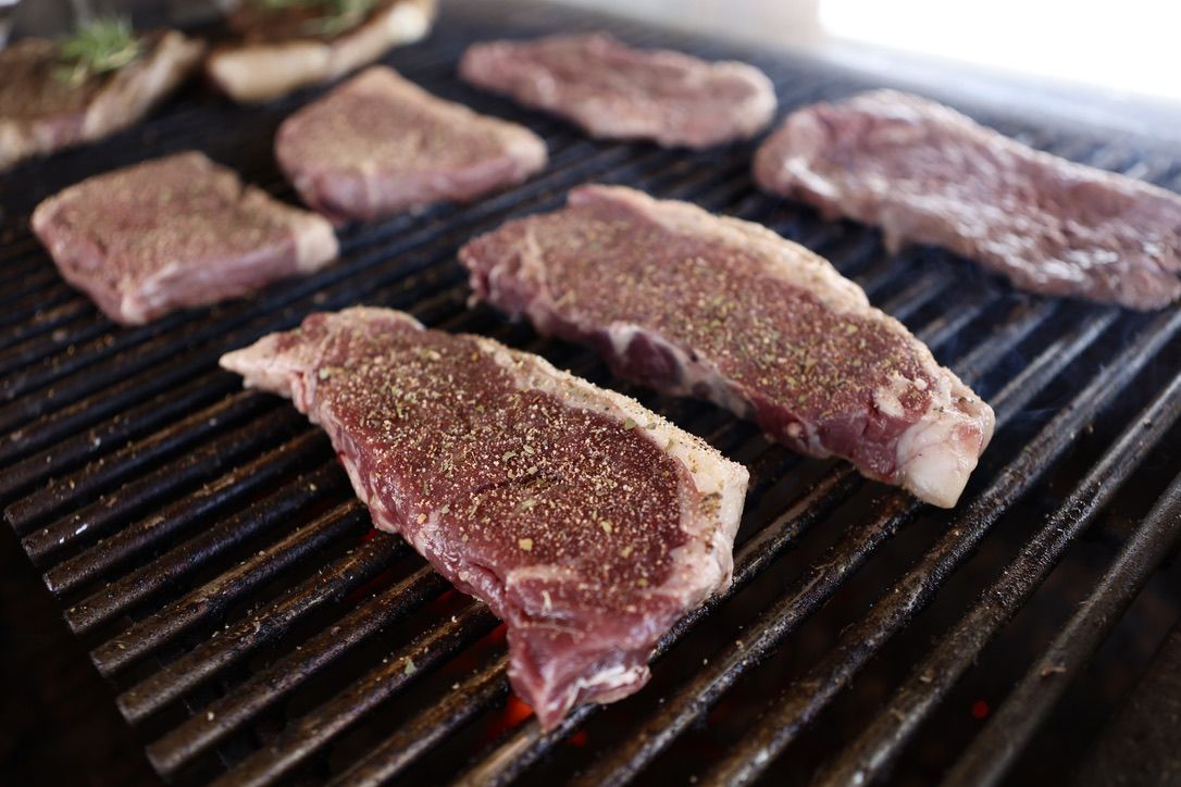Thick-cut seasoned steaks cooking on a grill with visible smoke, capturing the rich texture and flavor of Drummond Ranch’s premium cuts.
