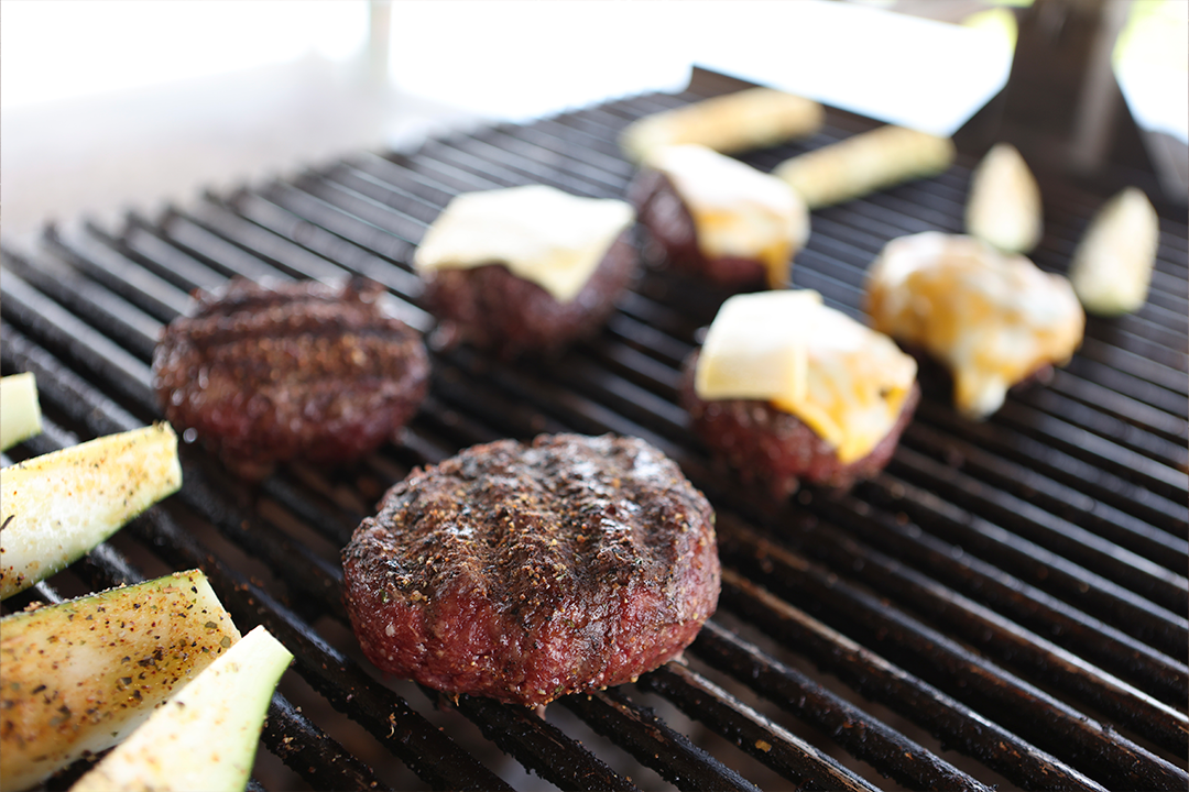 Seasoned beef patties and sliced vegetables grilling over open heat, with some burgers topped with melted cheese.