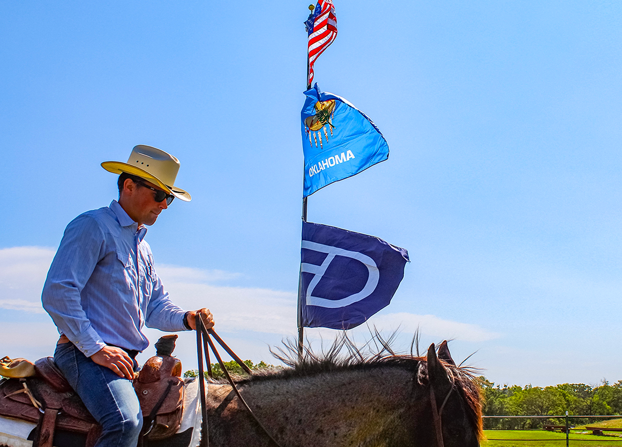 A cowboy in a hat and sunglasses rides horseback beneath waving American, Oklahoma, and Drummond Ranch flags on a clear, sunny day—capturing ranch pride and Western tradition.