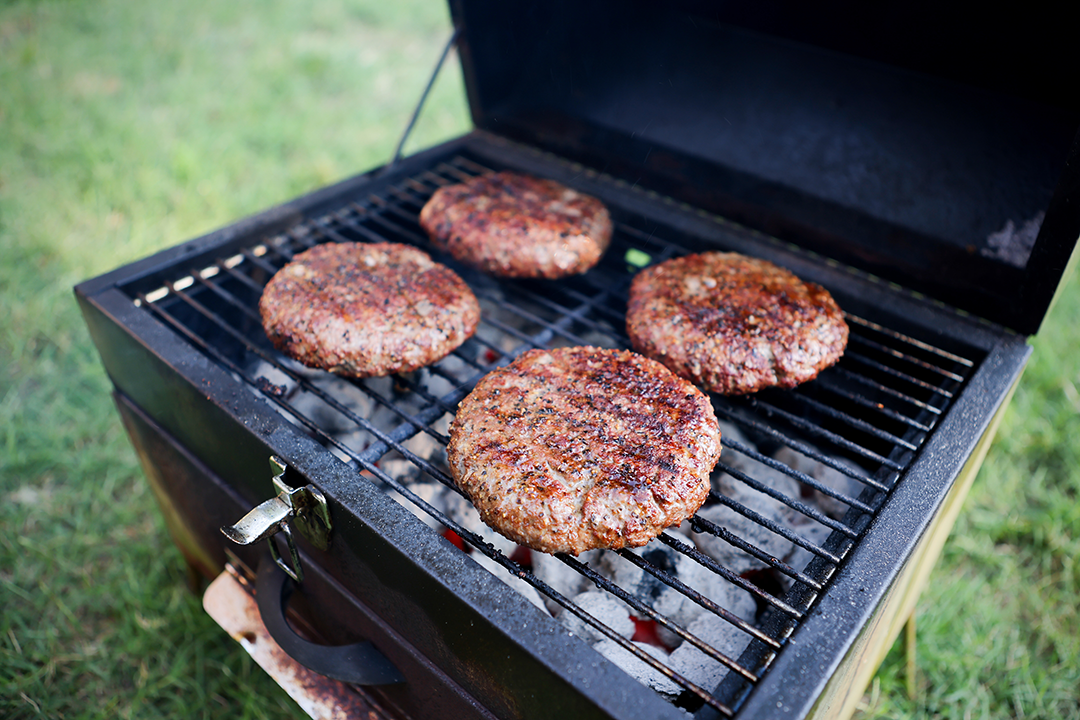 Four well-seasoned hamburger patties cooking over hot coals in a charcoal grill, ready to serve with that classic backyard barbecue flavor.