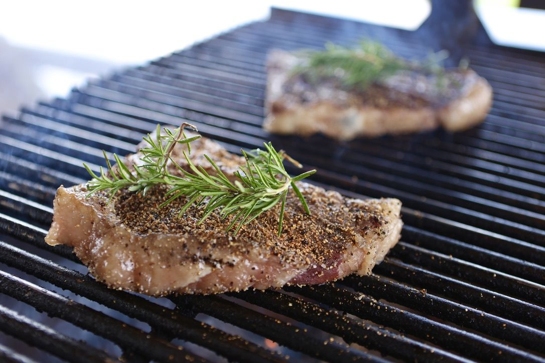 A close-up of a seasoned steak topped with fresh rosemary on a grill, with another steak cooking in the background—highlighting the savory preparation of Drummond Ranch beef.