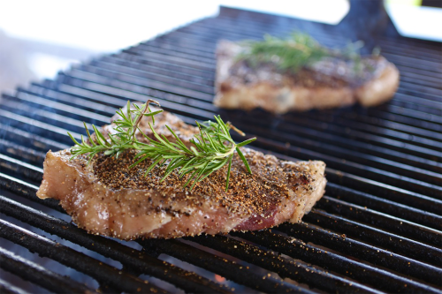 Seasoned steak with a sprig of rosemary cooking on a grill grate, with another steak blurred in the background.