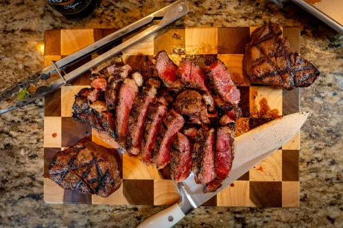 Overhead view of sliced medium-rare grilled steak on a checkered wooden cutting board with a chef’s knife and tongs on a granite countertop.