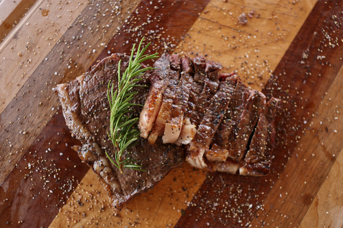 Sliced grilled steak topped with a sprig of rosemary on a wooden cutting board, seasoned with coarse salt and cracked pepper.